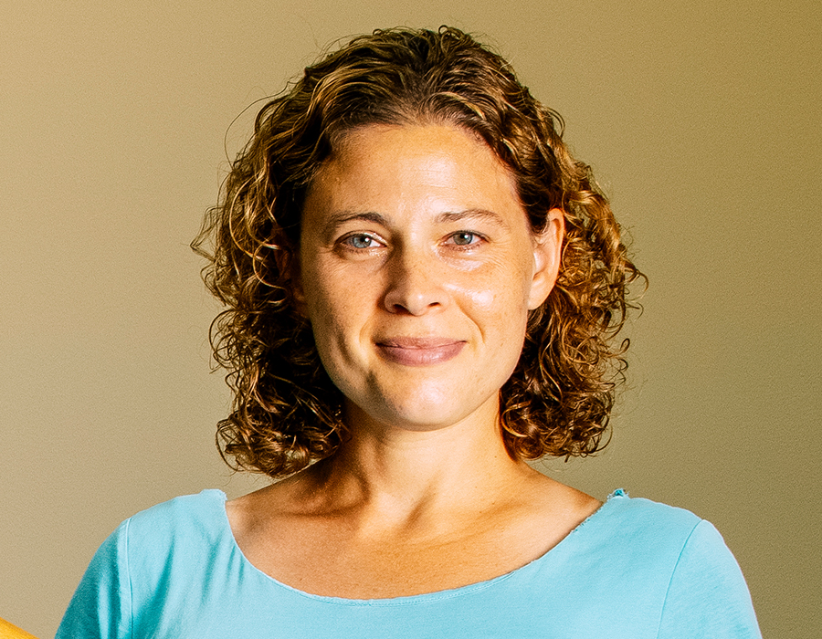 A woman with brown curly hair stands in front of a blank wall.