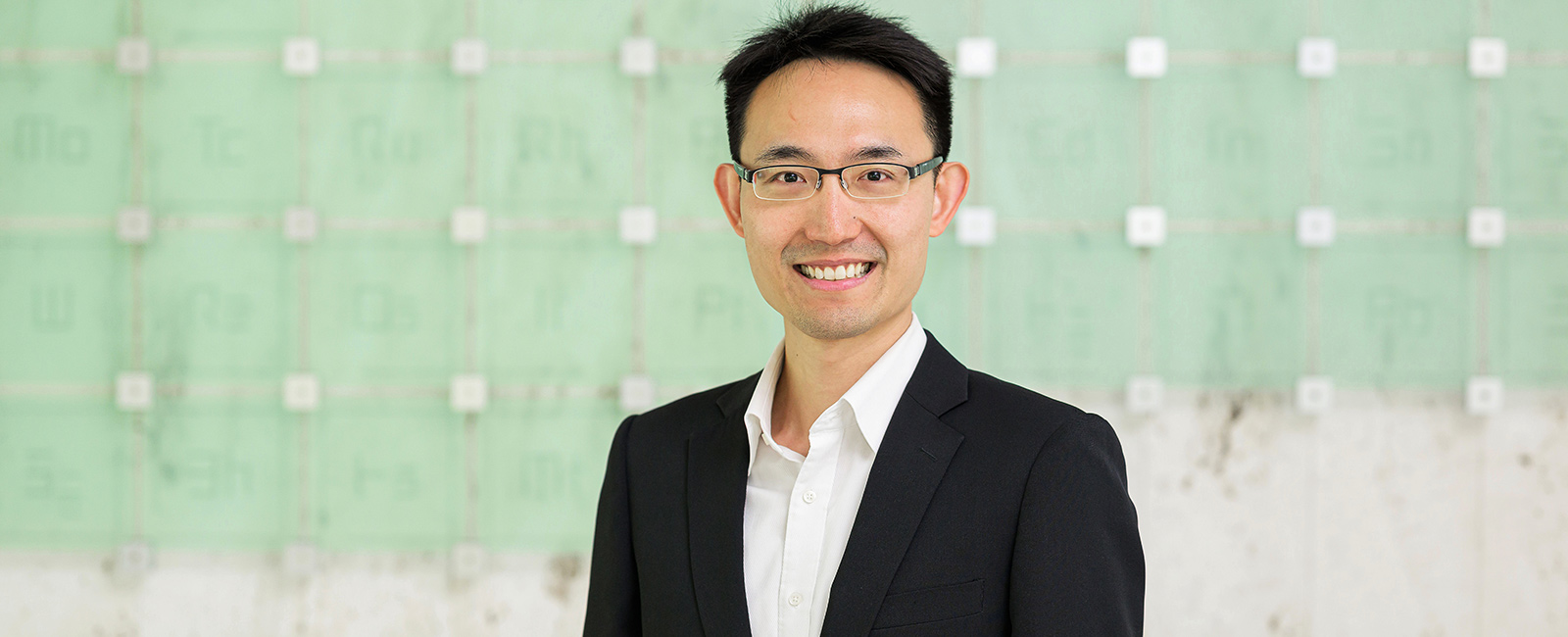 A man smiles in front of a green glass periodic table sculpture.