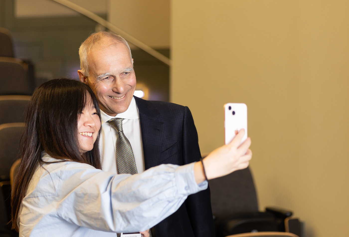 A student with long black hair holds her phone at arm's length to capture a photo of herself with Moungi Bawendi.