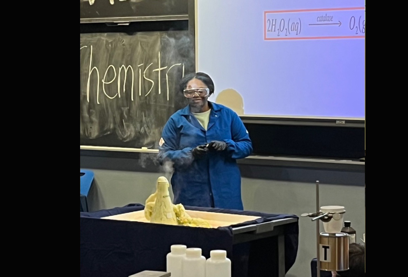 Michaela wears a lab coat and eye protection and stands behind a glass tube filled with white foam.