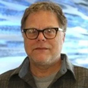 Art Spivack wears black plastic glasses and stands in front of ocean water.