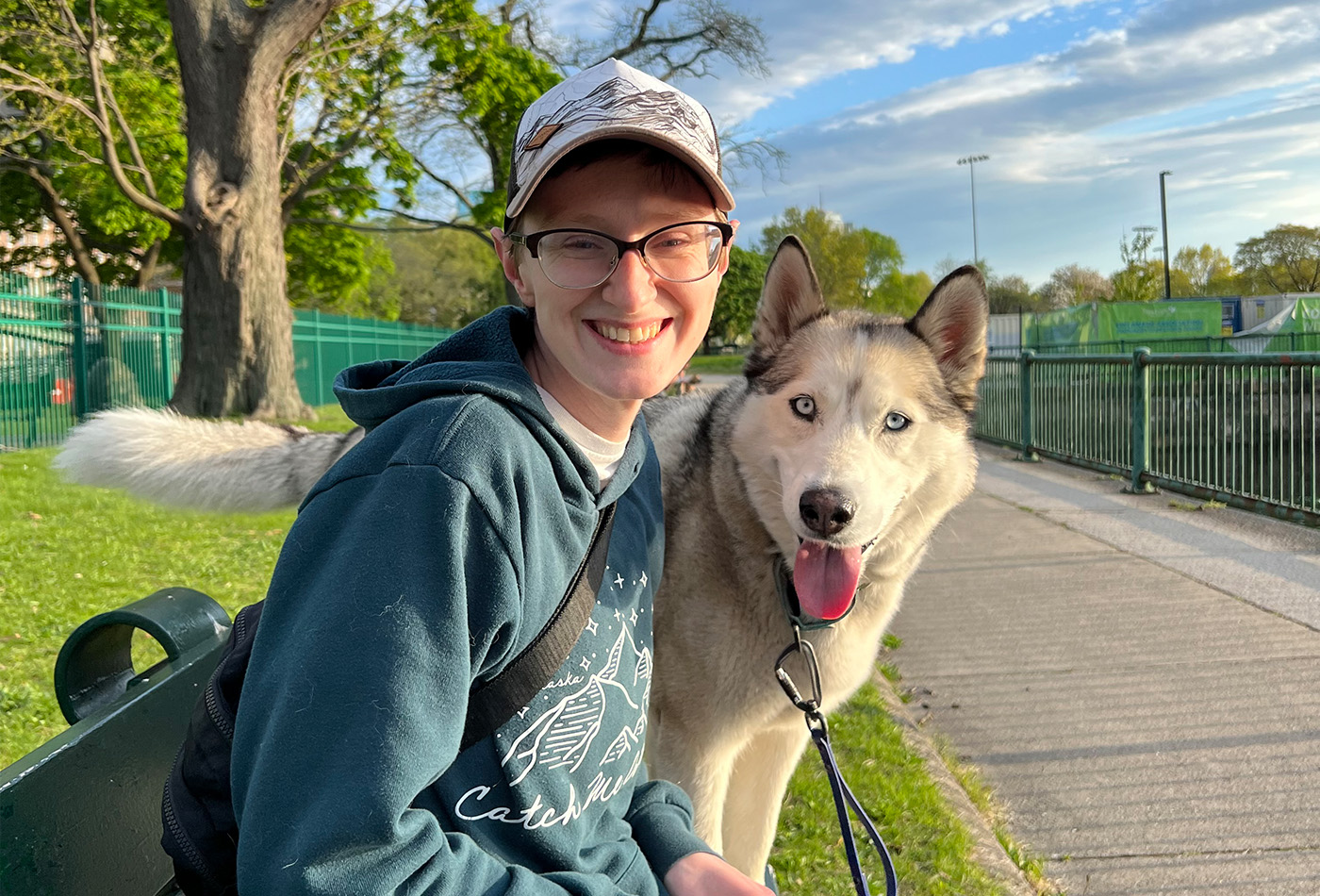 Beck Hanscam smiles on a bench beside a white husky with its tongue out.